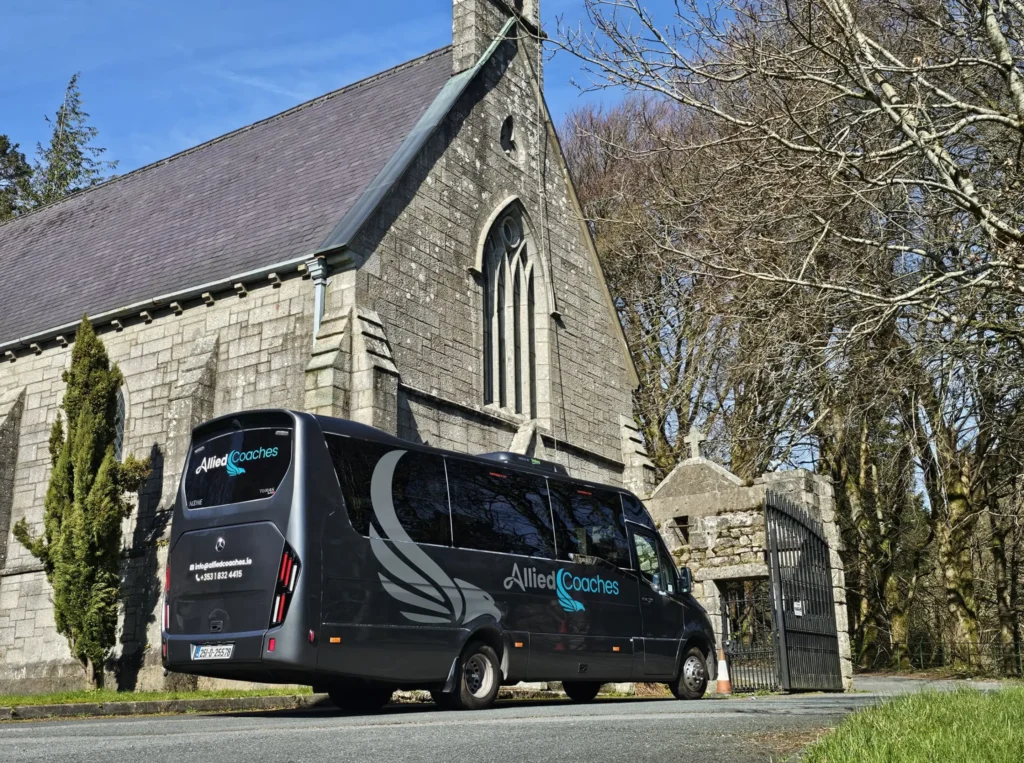 Black Allied Coaches van parked on a slight incline beside a stone church with arched windows and bare trees under a blue sky.