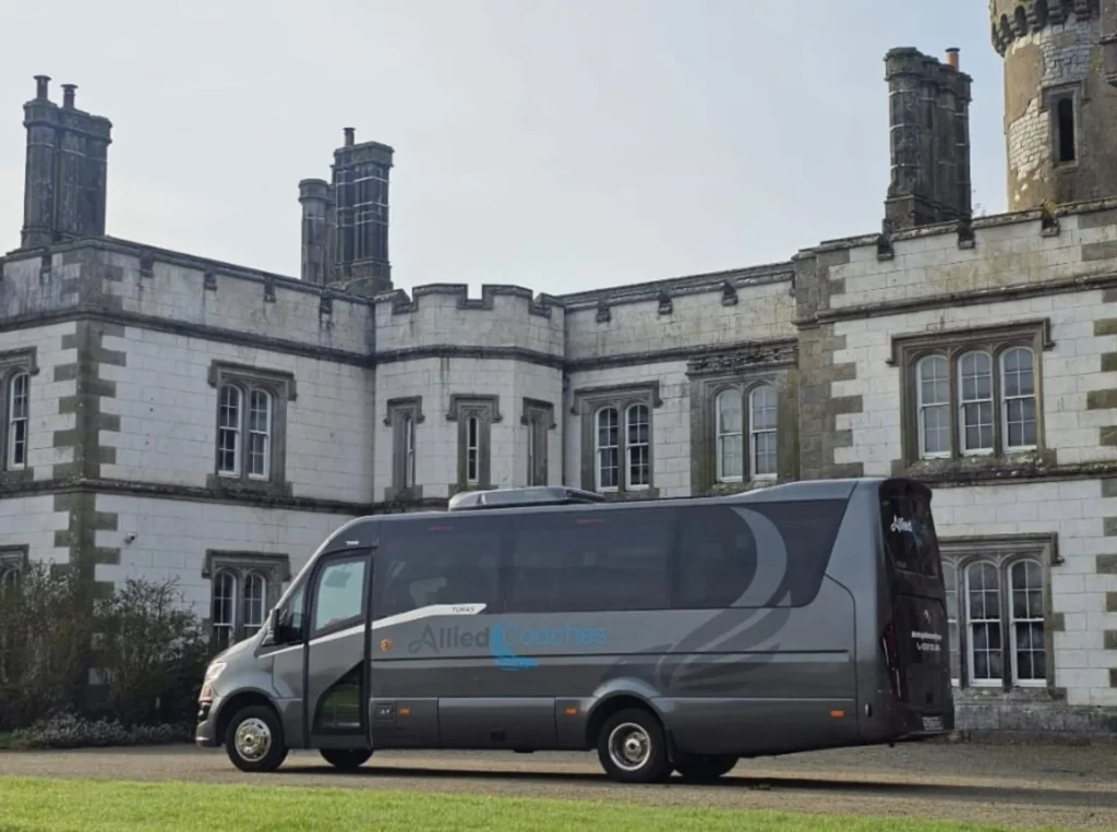 Grey coach bus parked in front of a historic stone building with tall chimneys and battlement-style walls.