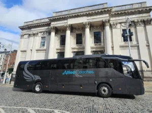 Black coach bus with Allied Coaches logo in blue parked in front of a neoclassical stone building with tall columns and ornate details under a blue sky.